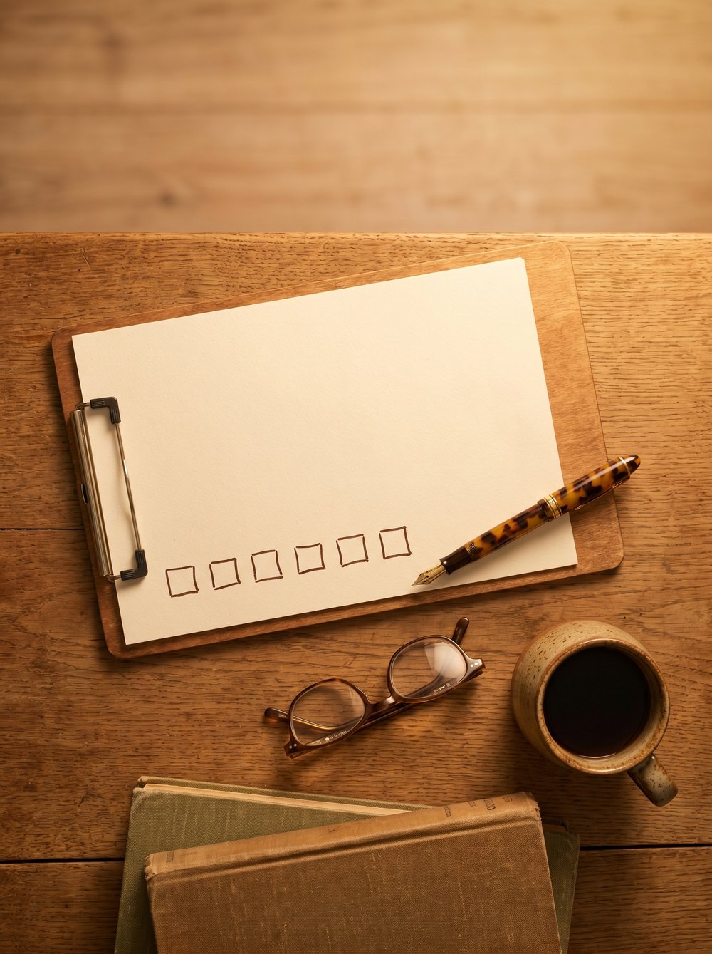 A clipboard with a blank check-list page, fountain pen, reading glasses and coffee on a warm wooden desk.